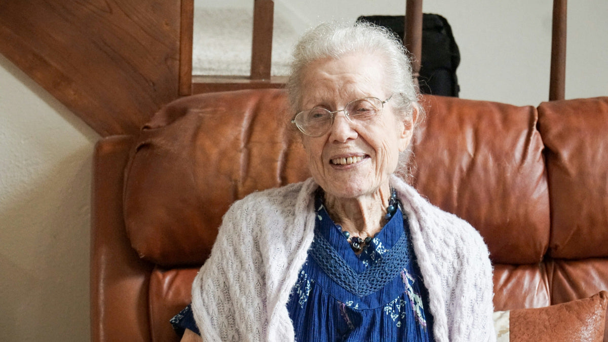 Femme âgée souriante portant des lunettes, un cardigan clair et un collier bleu Imagen